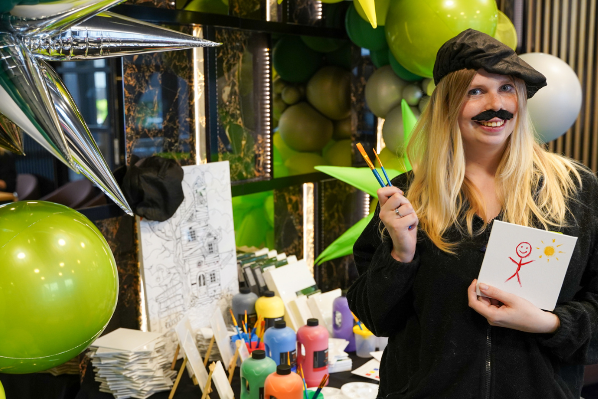 woman smiles wearing an artists beret and a fake moustache holding a paintbrush