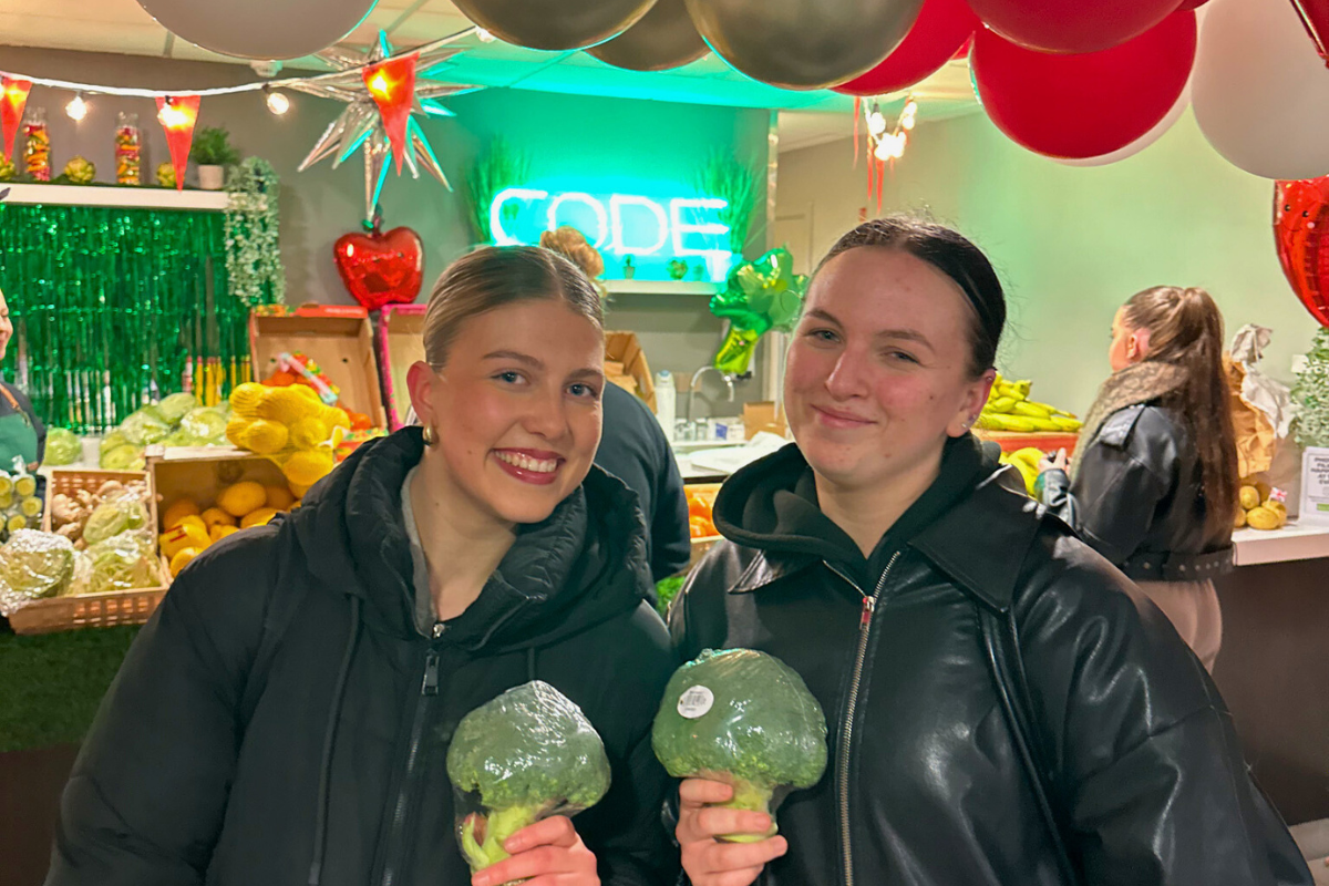 Two girls posing in front of the fruit and veg stall with broccolis in their hands