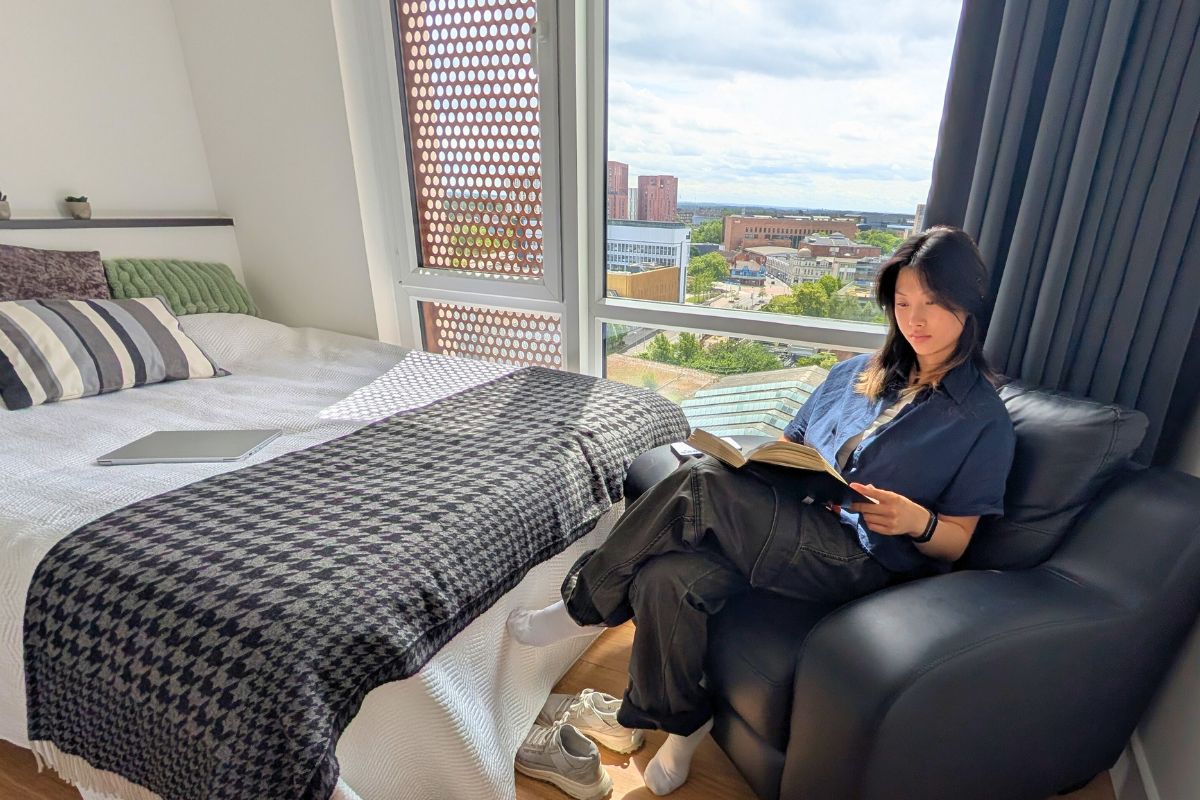 A student sitting in an armchair, reading a book in her room at CODE Student Accommodation.
