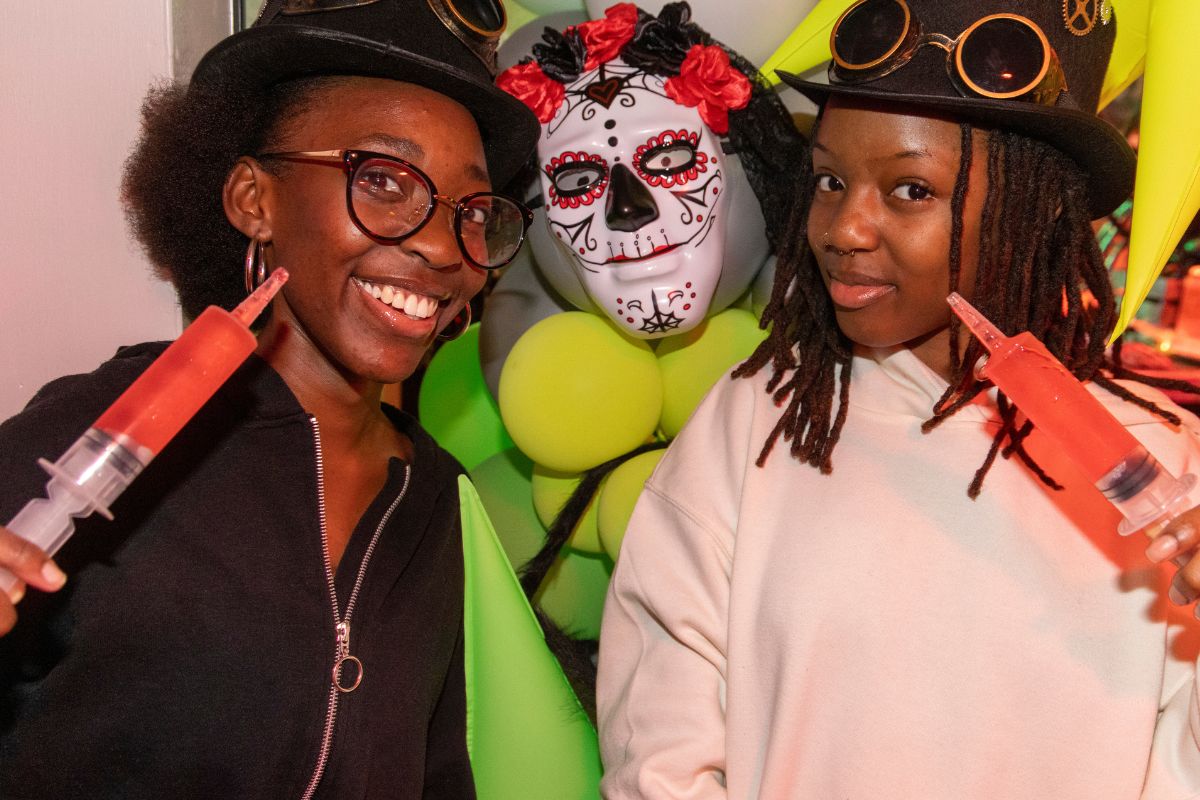Two CODE students in fancy dress posing with the syringe shots and the halloween decorations